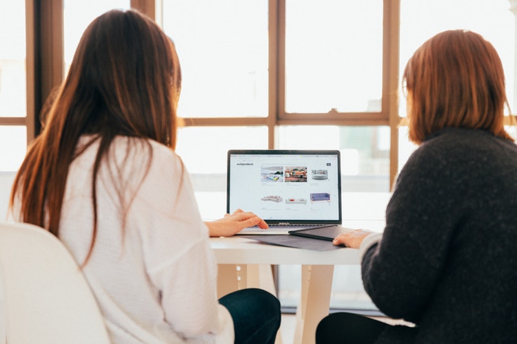 deux femmes à un bureau effectuent une recherche sur un ordinateur portable Etude-60-pourcent-des-visites-sur-un-site-du-Tourisme-s-effectuent-depuis-un-moteur-de-recherches